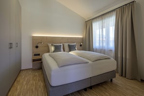Un dormitorio moderno con cabecera de madera, una cama grande con ropa blanca de cama, dos lámparas de noche y una ventana con cortinas translúcidas.