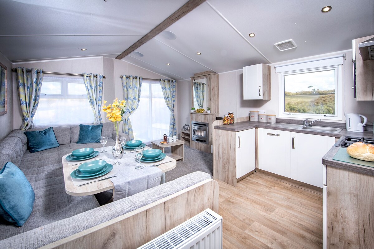 A modern RV interior with a dining area, kitchenette, and a large window offering a view of the outdoors.