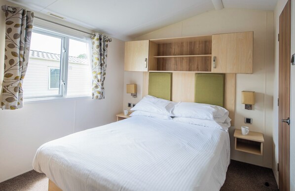 A bedroom with a bed, a window with curtains, a bedside table with a lamp, and built-in wooden cabinets above the bed.
