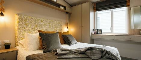 Un dormitorio con cama, mesitas de noche, una ventana con persianas y estantes empotrados con cuadros enmarcados.