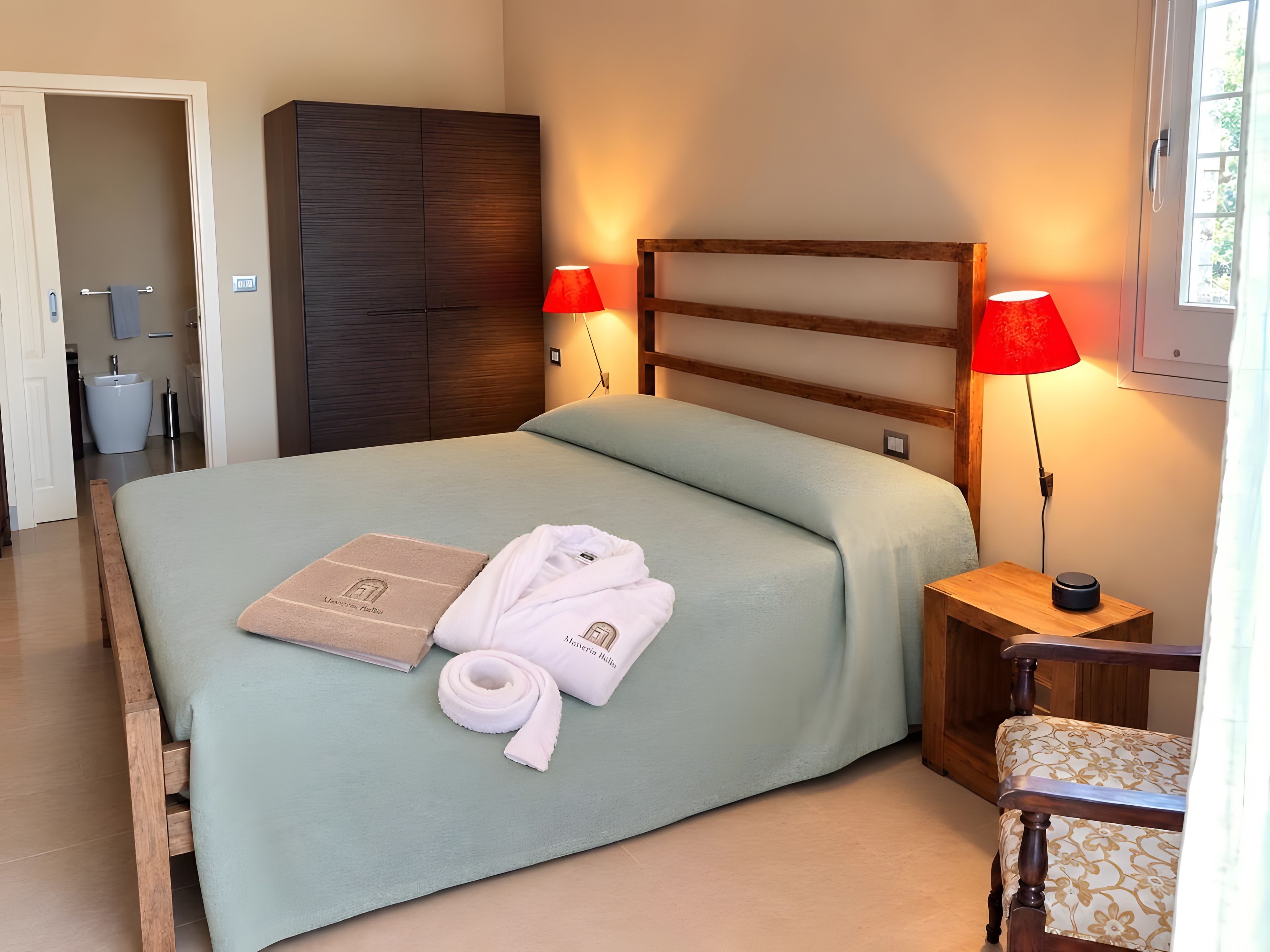 A neatly made bed with a light-colored bedspread, a wooden headboard, and two bedside lamps with red shades. There is a wooden nightstand with a lamp and a small table beside the bed.