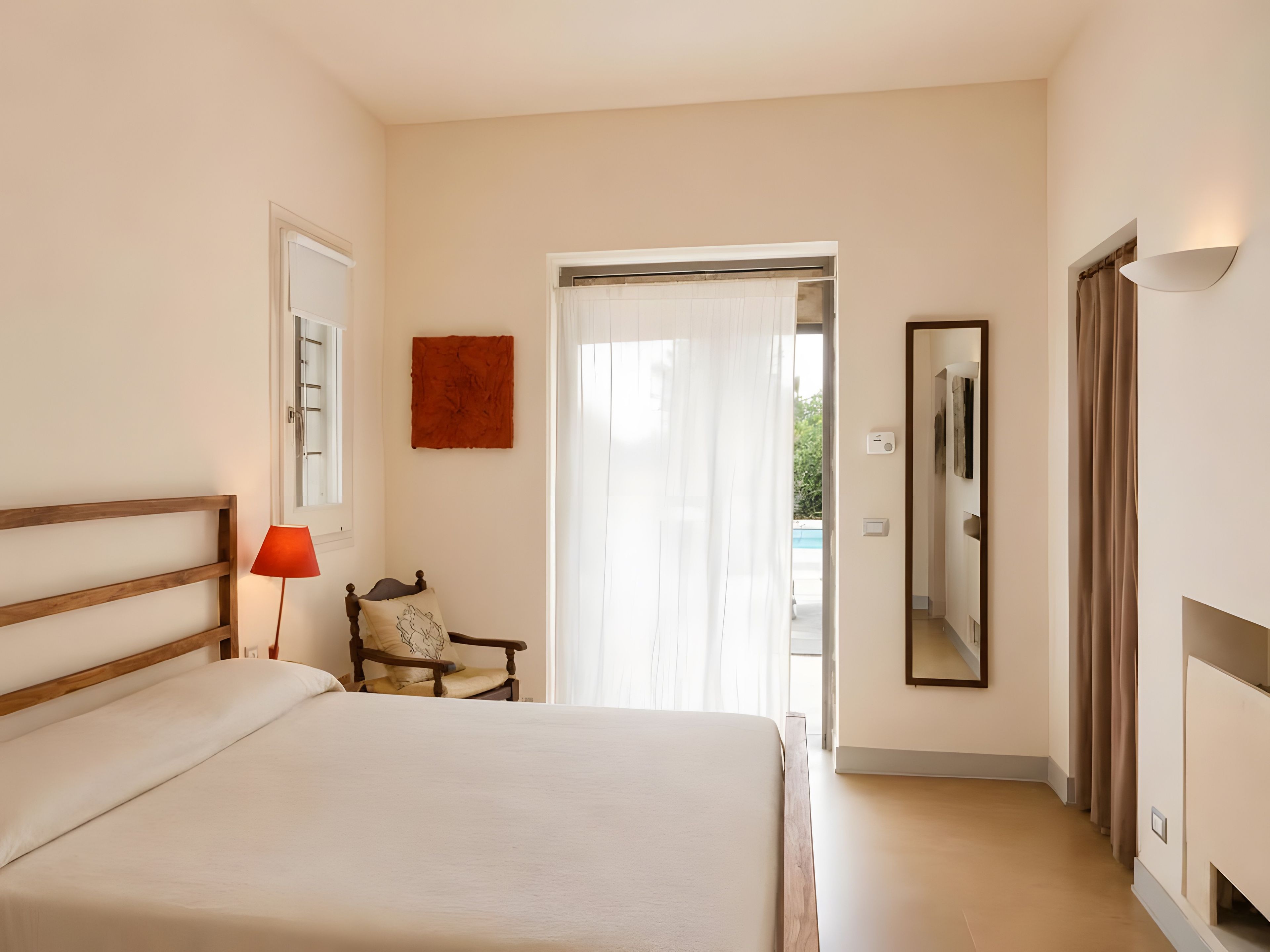 A hotel room with a bed, a chair, a lamp, a mirror, and a door leading to a balcony.