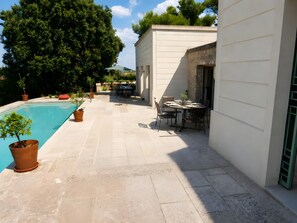 Zona de piscina con mesa para dos, rodeada de plantas en macetas y un muro de piedra.