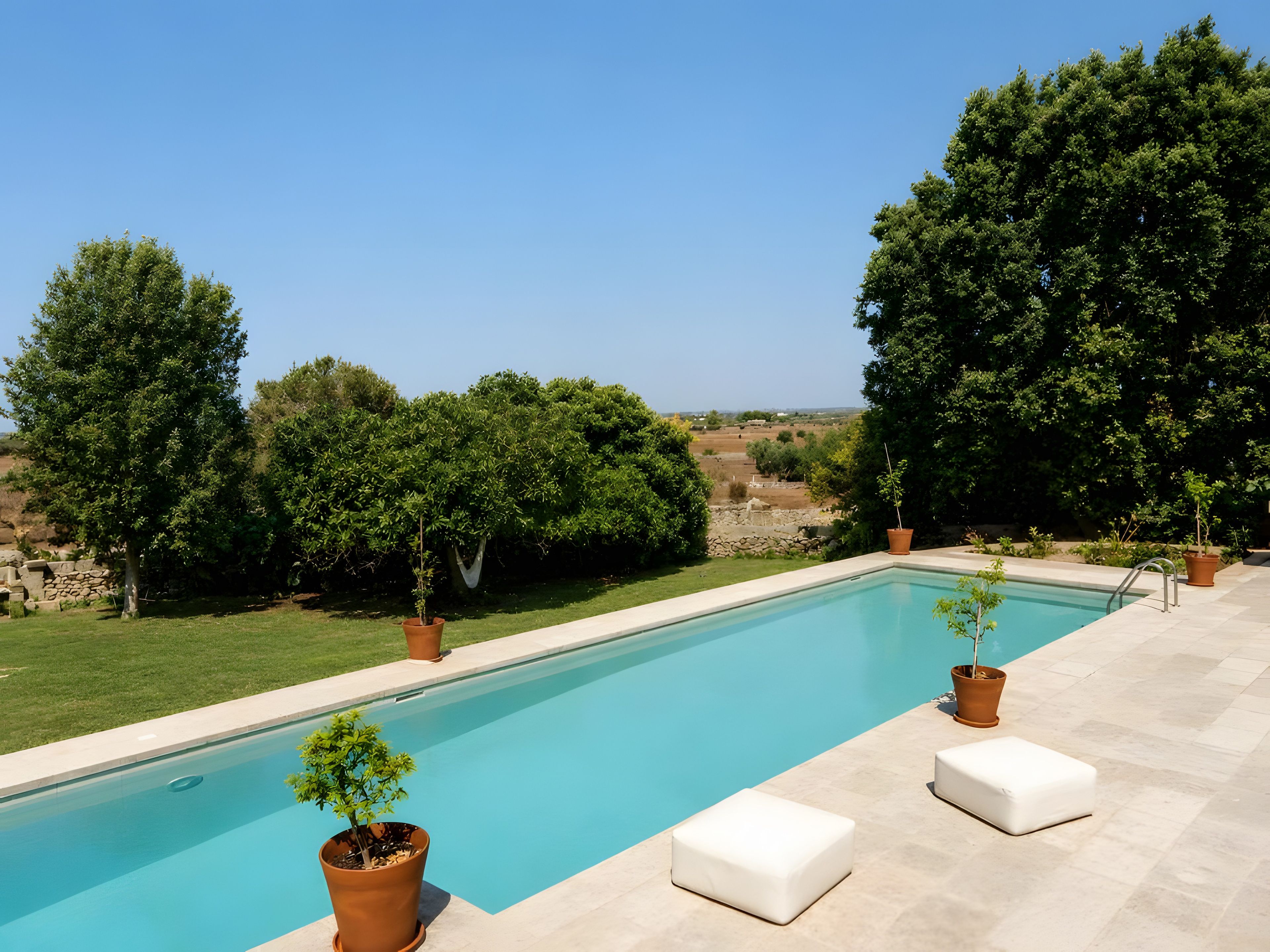 A rectangular swimming pool with a clear blue water surface, surrounded by a paved deck. There are two white cushioned seating areas and several potted plants. The background features a natural landscape with trees and a stone wall.