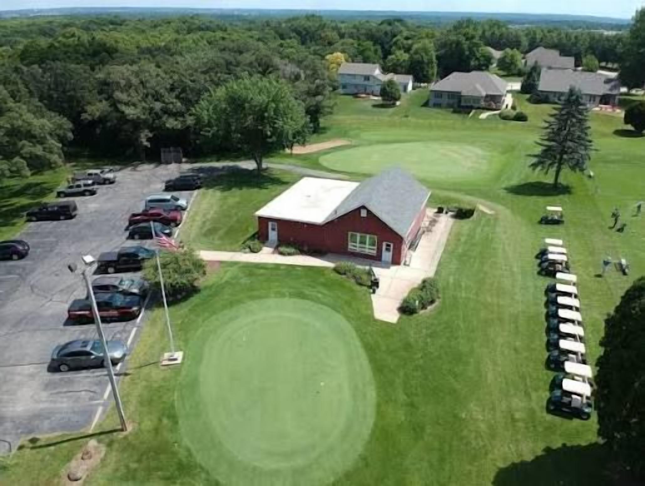 A golf course with a clubhouse, parking lot, and surrounding trees.