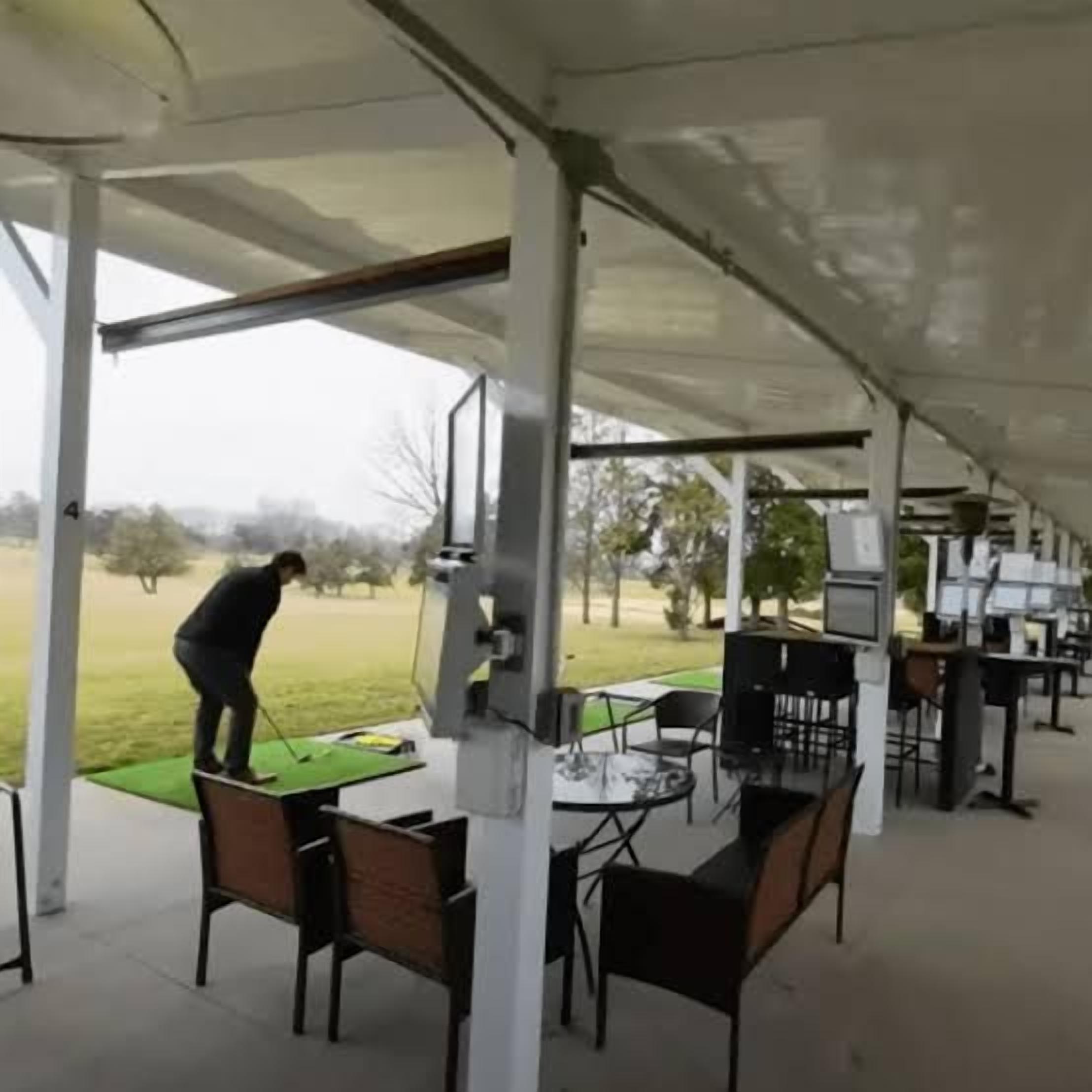 A covered outdoor area with tables and chairs, a green mat, and a person playing golf.