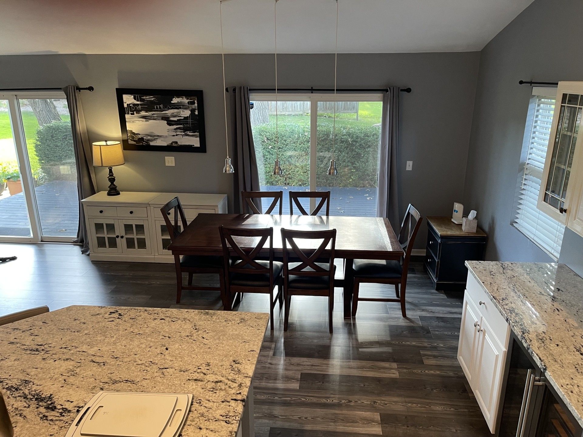 A modern kitchen with a granite countertop, a dining area with a wooden table and chairs, and a view of the outdoors through large windows.