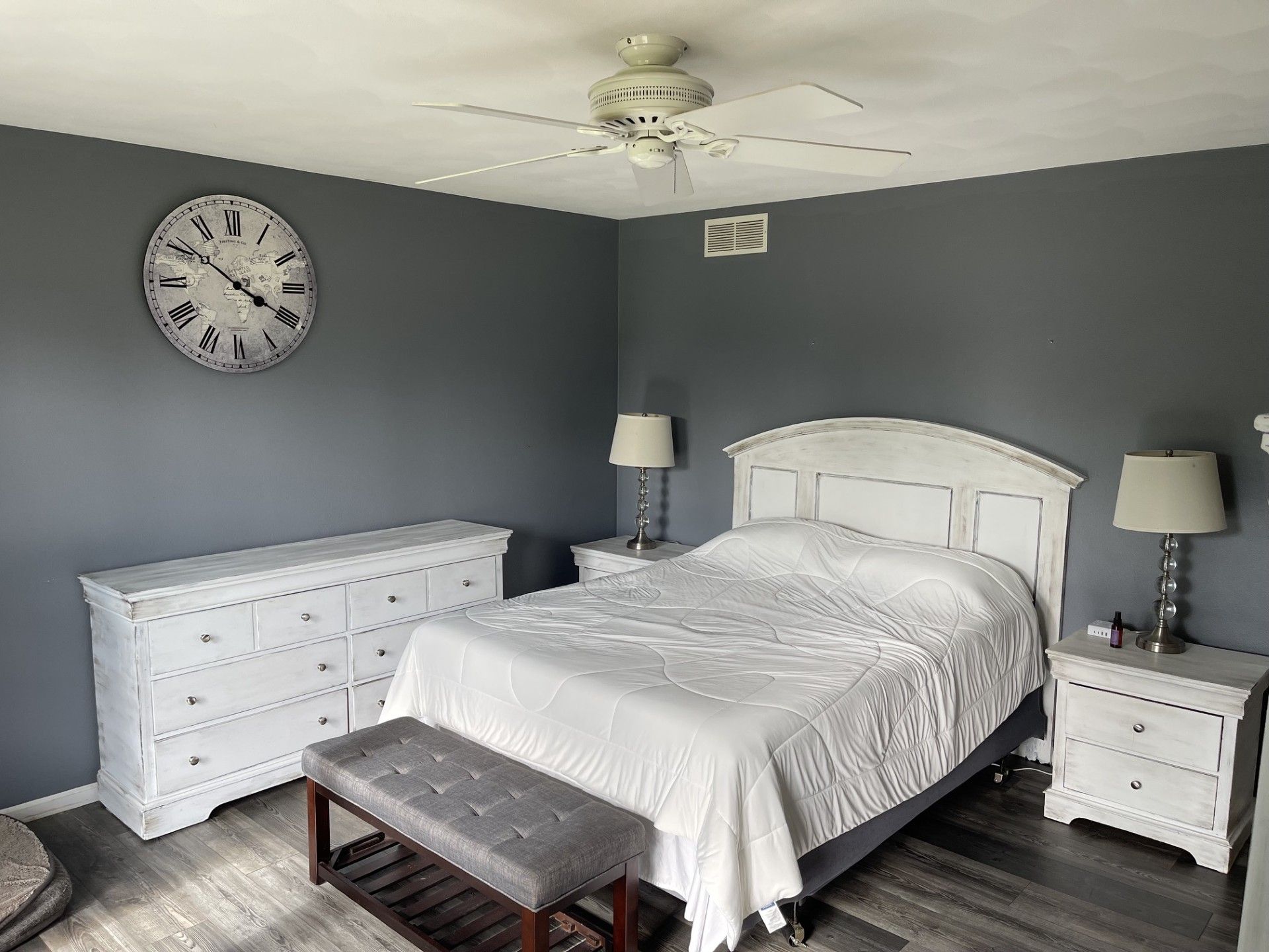 A bedroom with a white bed, a dresser, two nightstands with lamps, a bench, a ceiling fan, and a wall clock.