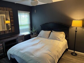 Ein Schlafzimmer mit Bett, Nachttischlampe, Fenster mit Jalousien und einem Spiegel an der Wand.