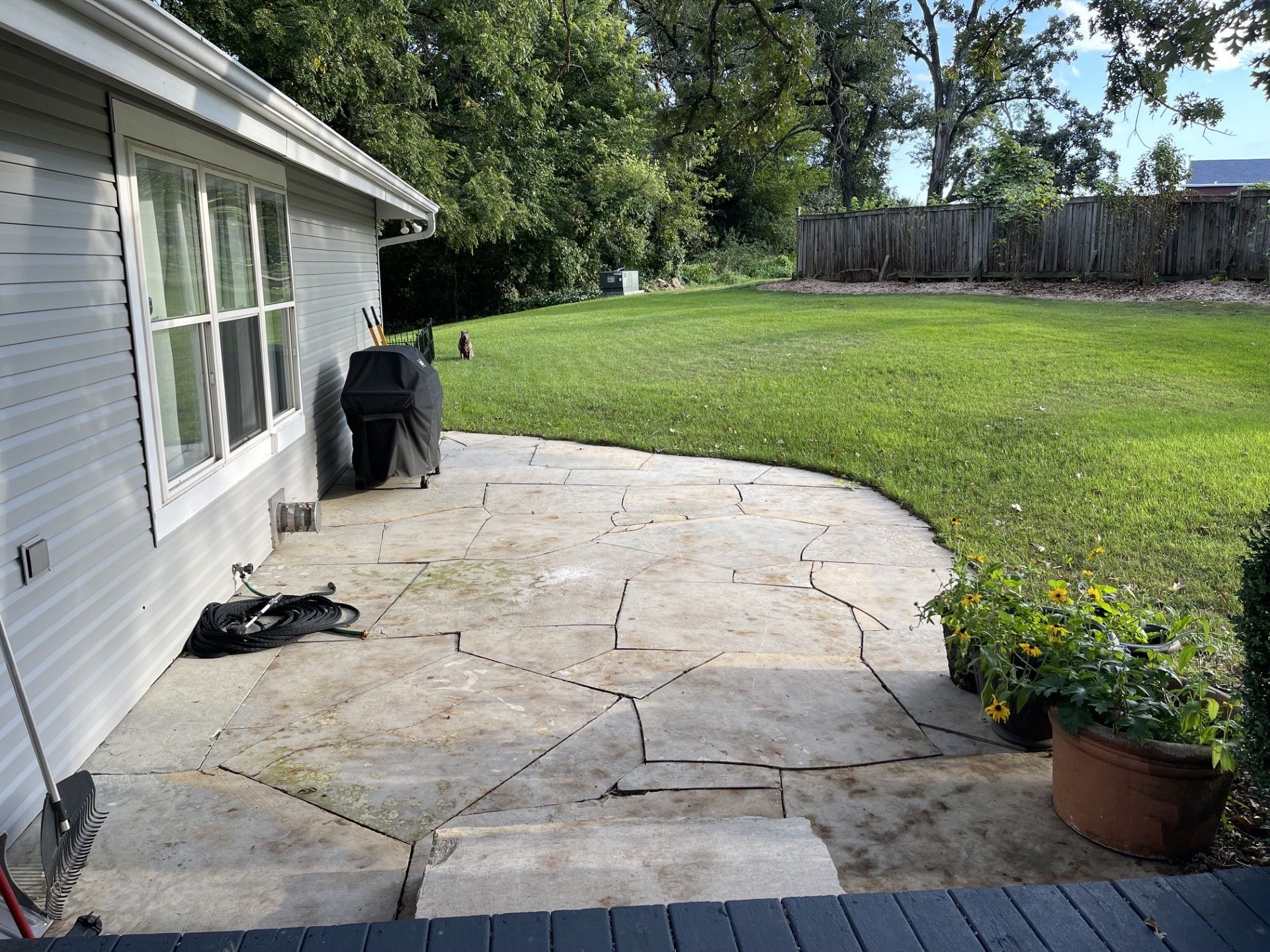 A residential backyard with a stone patio, a lawn, a wooden fence, and a house with a garage.