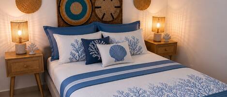 Una cama bien hecha con colcha azul y blanca, flanqueada por dos mesitas de noche con lámparas, y una pared decorada con piezas de arte circular tejidas.