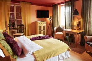 Quarto de hotel com cama, escrivaninha, cadeira, televisão e janela com cortinas.