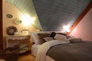 Quarto com teto inclinado, cama com várias almofadas, mesa de cabeceira com abajur e relógio de parede.