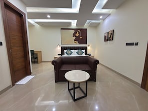 Quarto de hotel moderno com sofá de couro marrom, mesa redonda, cama com cabeceira e obras de arte nas paredes.