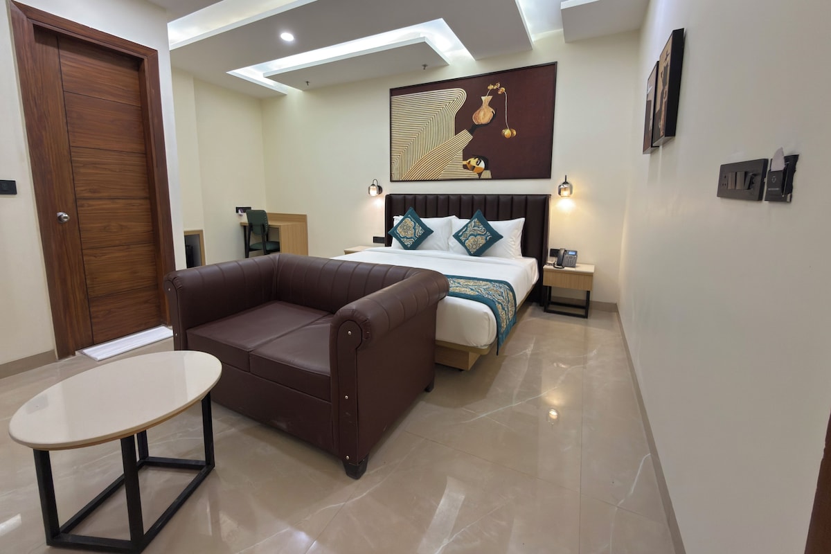 Quarto de hotel moderno com cama, sofá de couro marrom, mesa redonda pequena e obra de arte na parede.