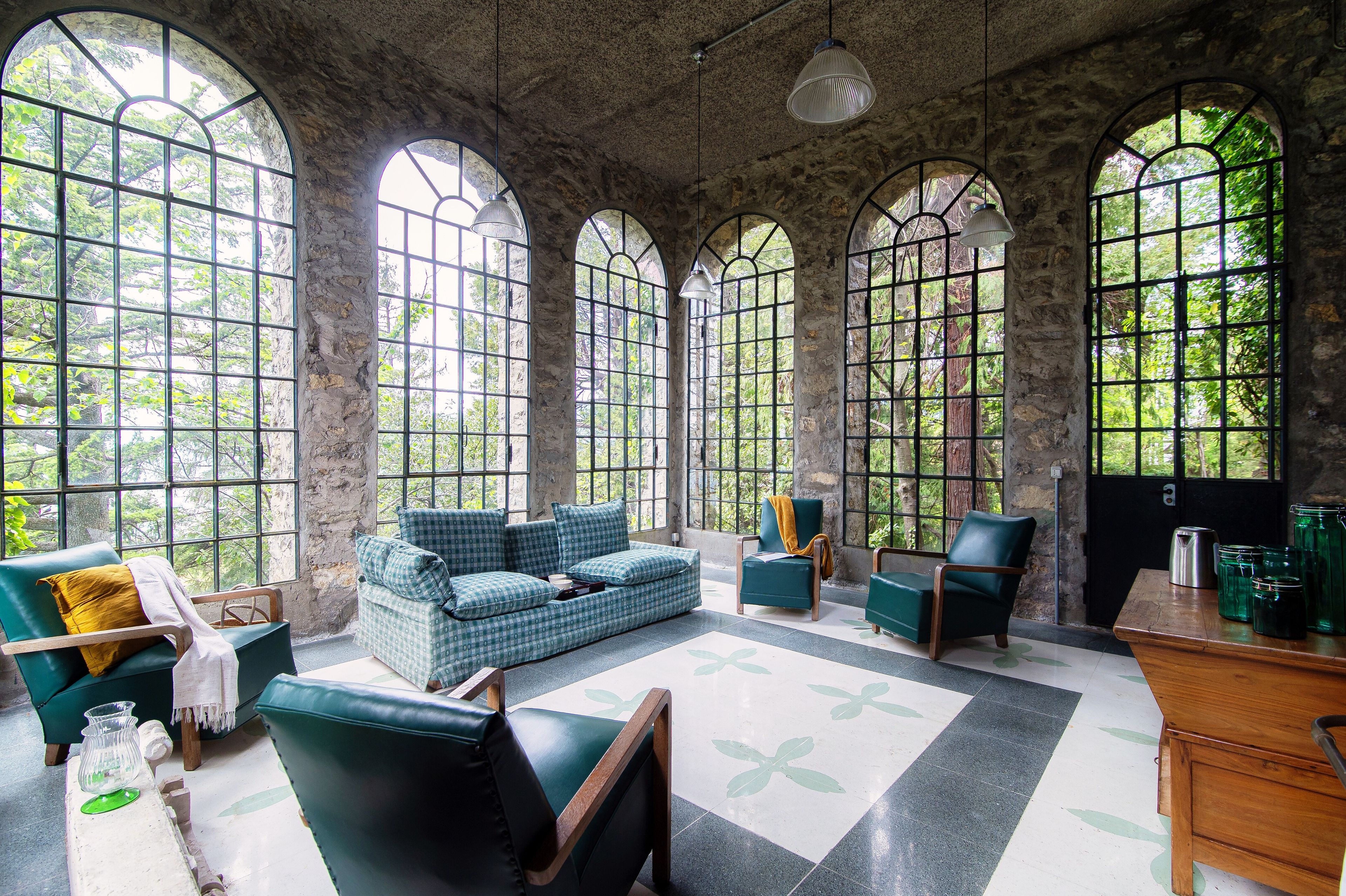Une pièce avec un mur en pierre, de grandes fenêtres en arc de cercle, un canapé vert, une chaise en bois et une petite table sur laquelle sont posés des pots.