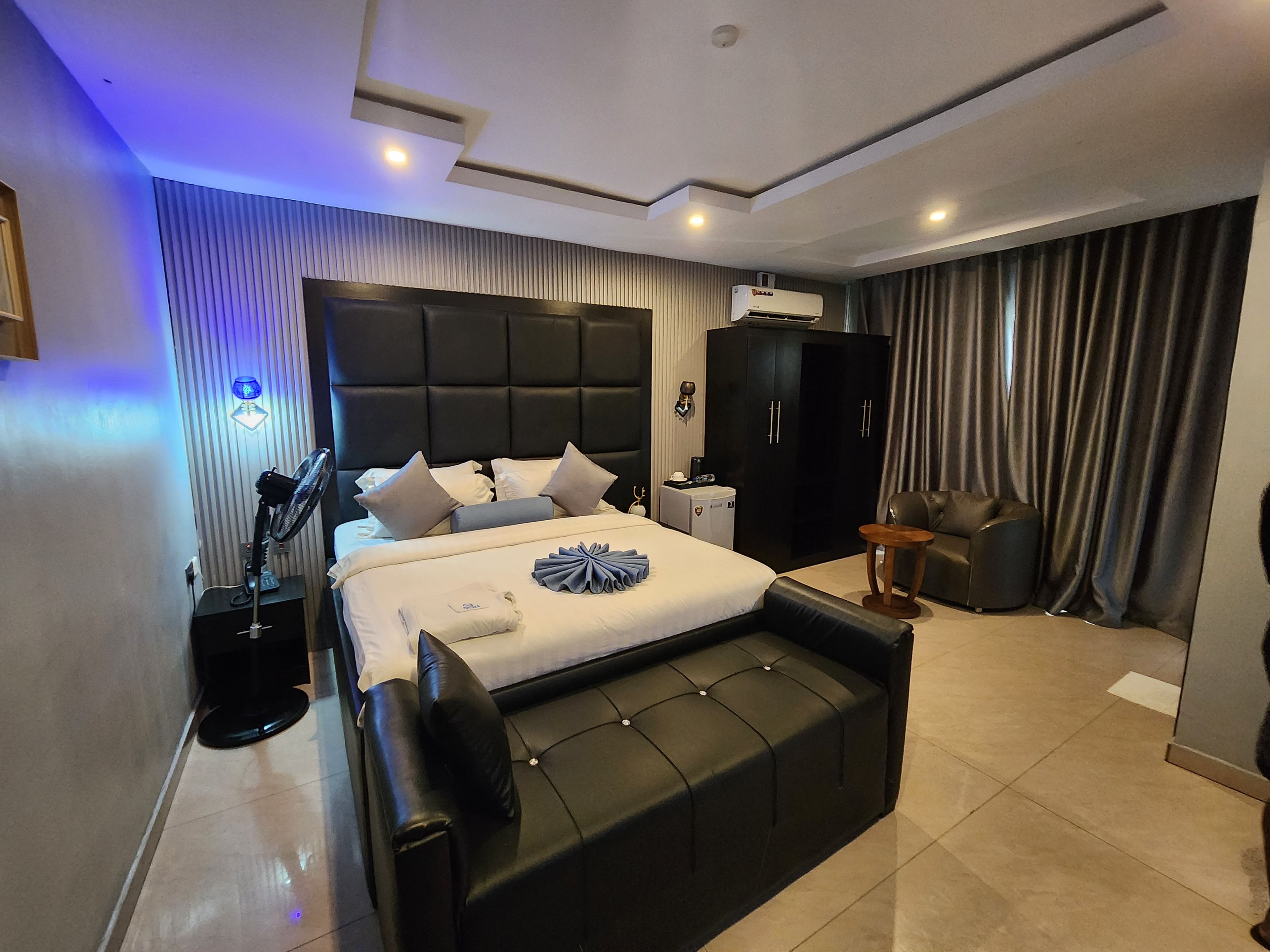 A modern bedroom with a large bed, a black leather sofa, an air conditioning unit, and a ceiling fan.
