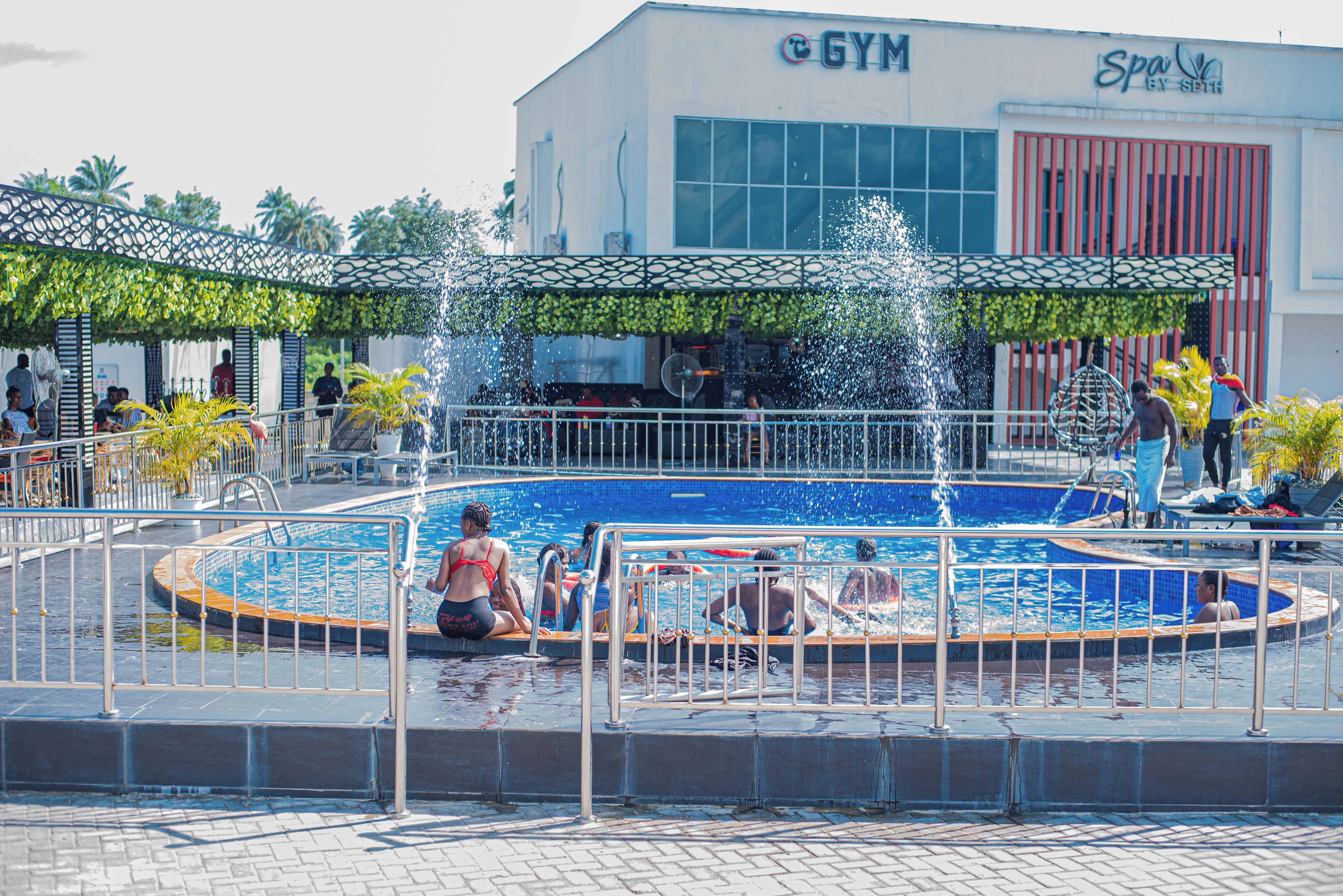 Ulkoilmauima-allas, jossa on suihulähde, ja sen ympärillä on metalliaita. Lähellä on rakennus, jonka kyltissä lukee: ”GYM SPA”.