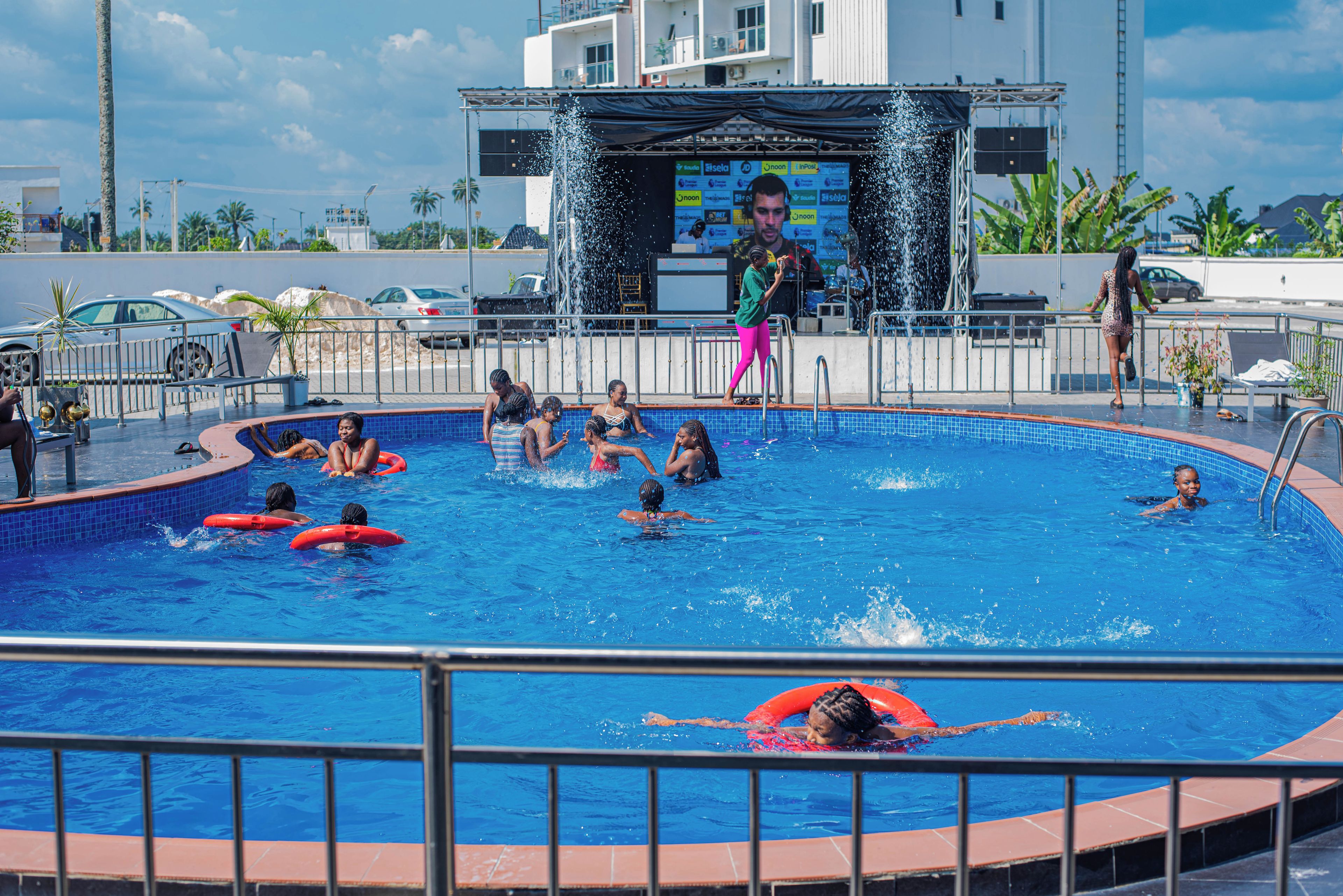 A swimming pool with multiple people, some on inflatable rings, and a stage with a screen in the background.