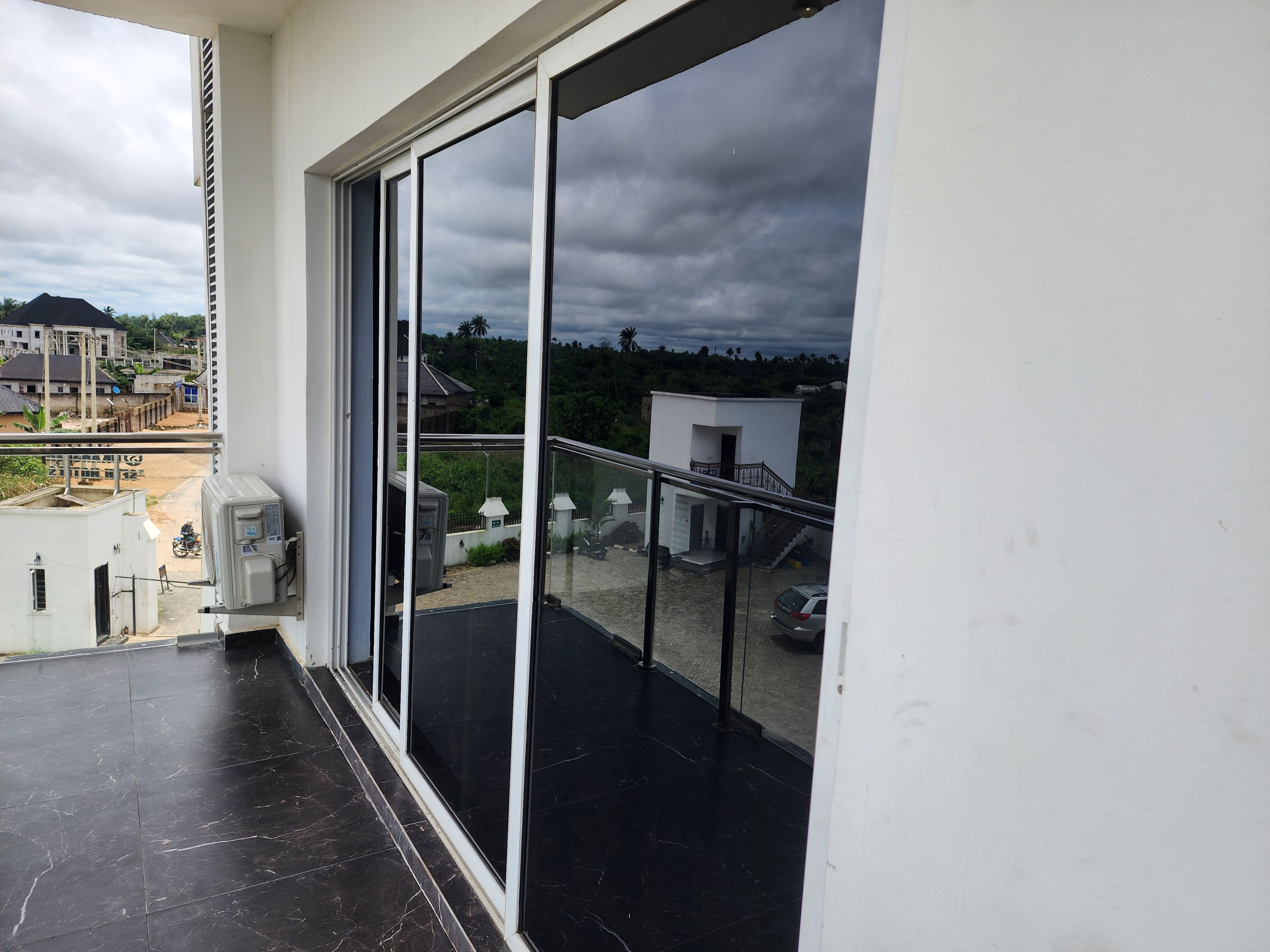 A balcony with a glass door leading to a terrace, a white wall, and a view of a residential area with houses and greenery.
