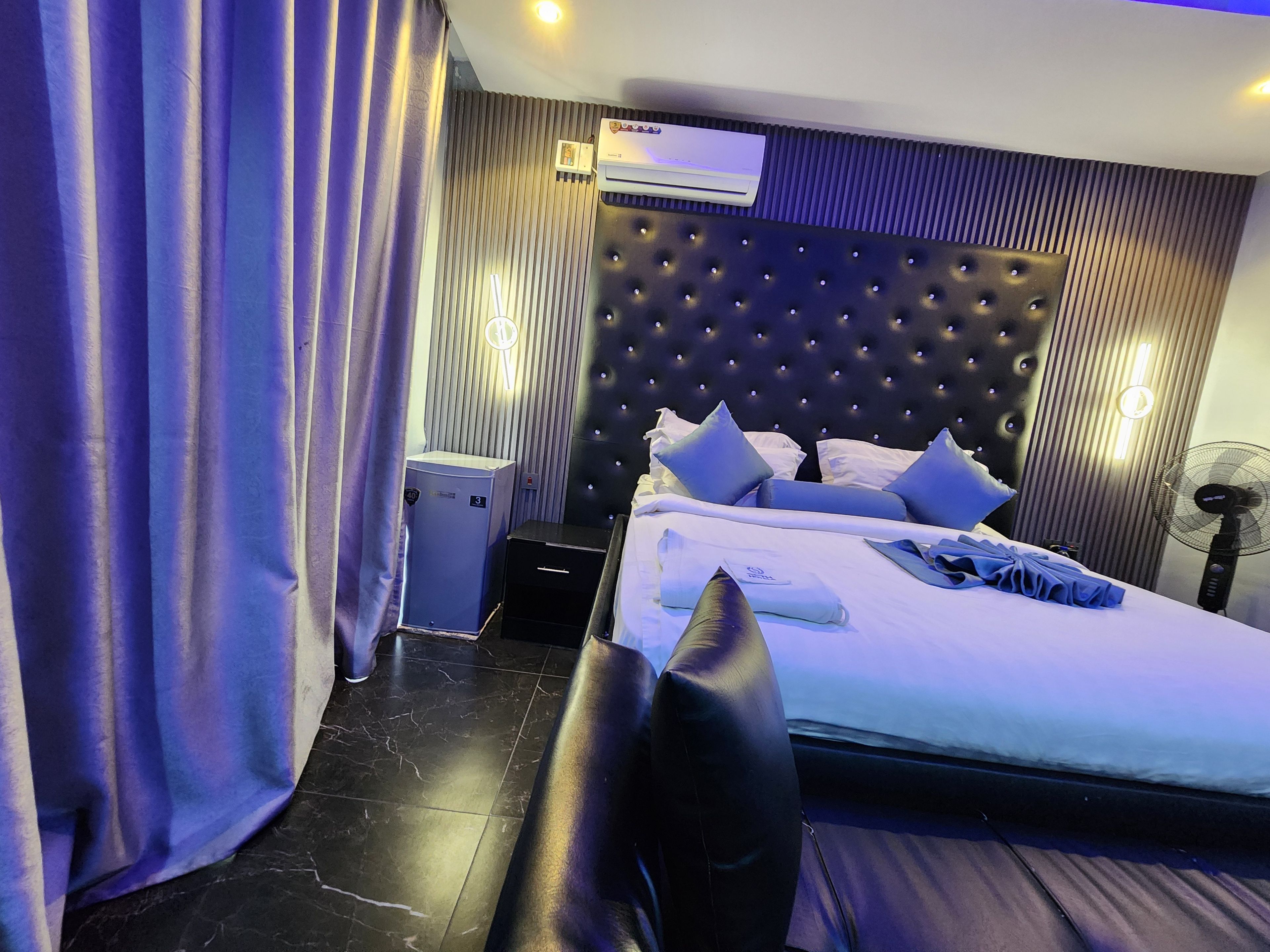 A hotel room with a bed, a nightstand, an air conditioning unit, a wall-mounted fan, and a curtain.
