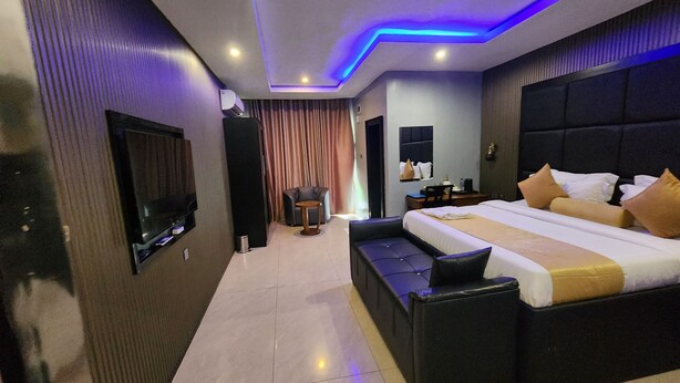 A modern hotel room with a large bed, a flat-screen TV mounted on the wall, a seating area with a small table, and a ceiling with blue LED lighting.