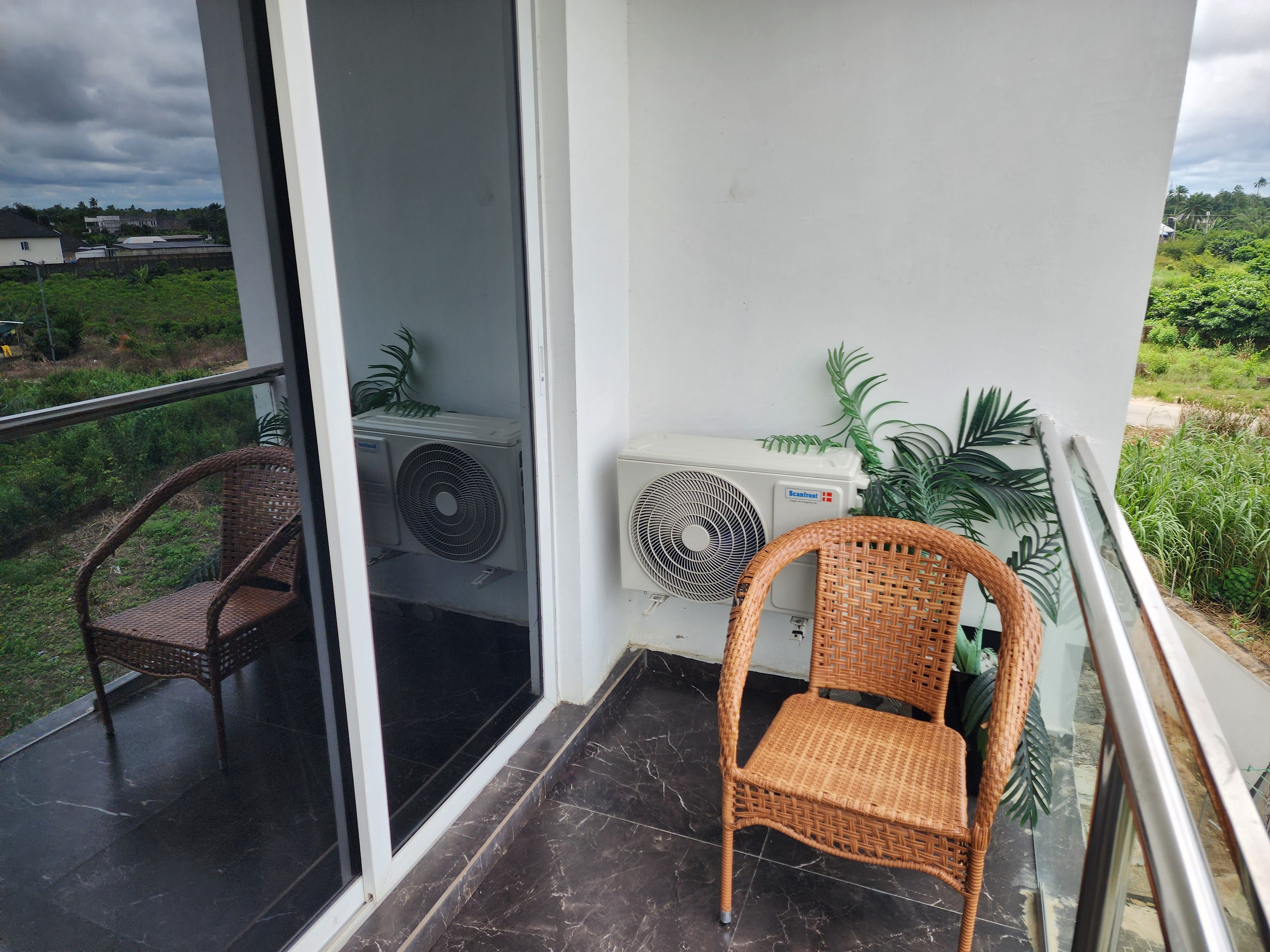 Un balcón con dos sillas de mimbre, un equipo de aire acondicionado y vista a la vegetación.