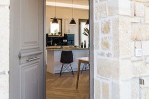 Ein offenes Durchgang führt in eine moderne Küche mit Pendelleuchten, einen Essbereich mit einem Holztisch und Stühlen sowie eine Steinwand.
