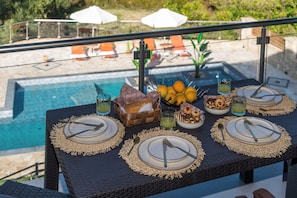 A dining table set for four with plates, cutlery, and a basket of bread and oranges, overlooking a pool area. - Villa Licaj - Four Bedroom Villa, Sleeps 8 (Tragaki)