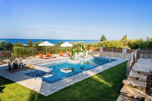 Ein Pool mit einer Steindeckplatte, umgeben von Gras und einer Steinmauer. Liegestühle und Sonnenschirme sind vorhanden.