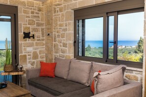 A room with a stone wall, a grey sofa with red cushions, and a view of the sea through large windows. - Villa Licaj - Four Bedroom Villa, Sleeps 8 (Tragaki)