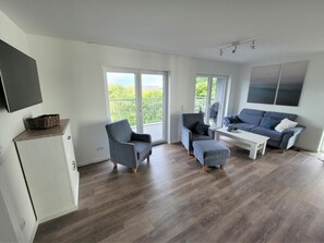 Ein modernes Wohnzimmer mit einer Couch, Sesseln, einem Couchtisch und einem an der Wand montierten Fernseher.