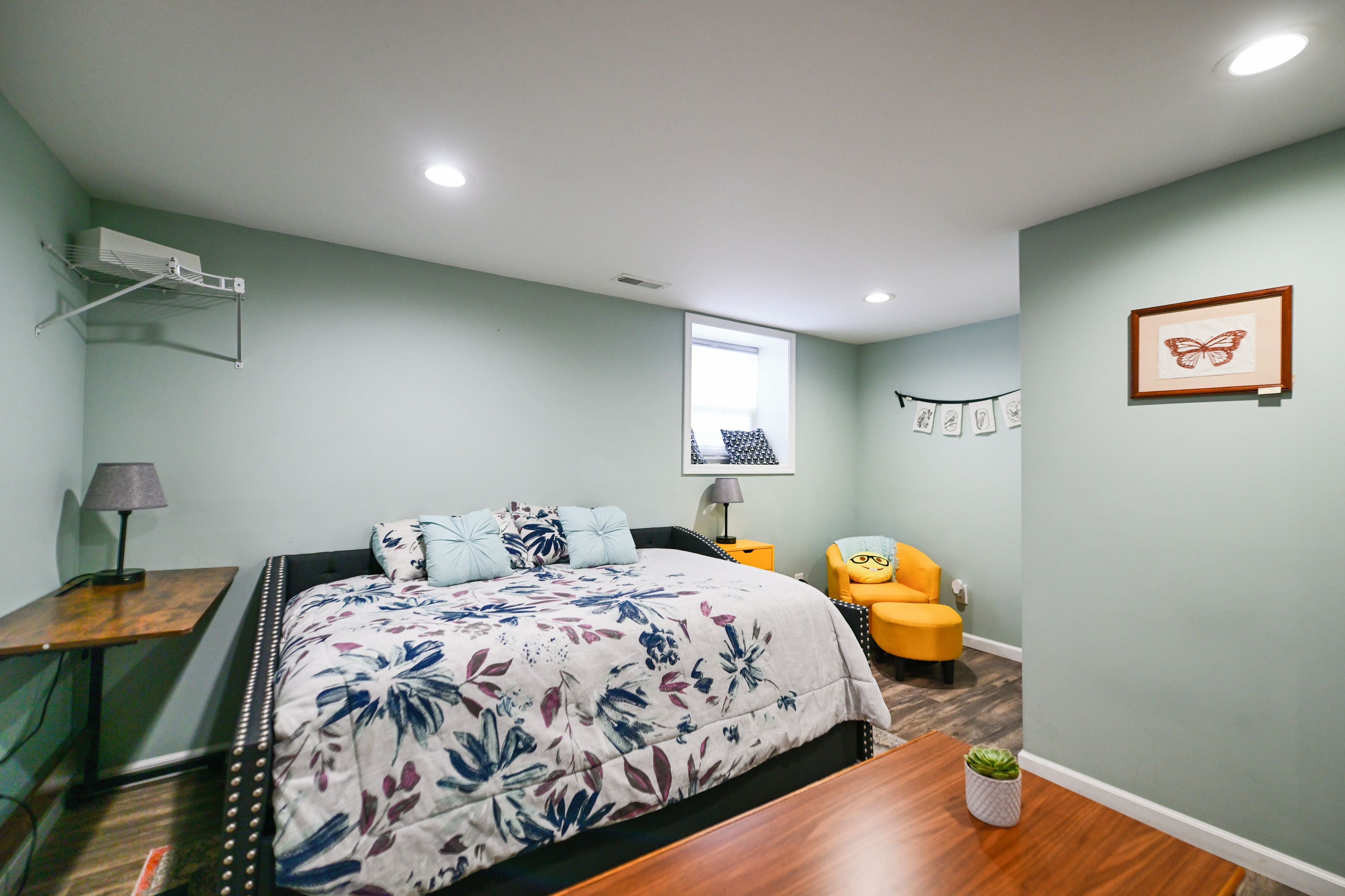 A bedroom with a bed, a desk, a chair, a window, and a wall-mounted shelf.