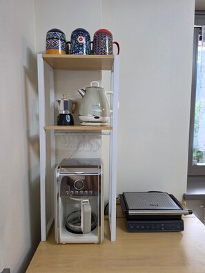Eine Kaffeemaschine, ein Wasserkocher und ein Laptop stehen auf einem Schreibtisch, über dem ein Regal mit drei Tassen angebracht ist.