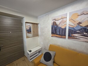 Ein Zimmer mit einem Holzschrank, einer weißen Bank und einem gelben Sofa mit einem schwarzen Kissen. An den Wänden hängen zwei Gemälde mit Berglandschaften.
