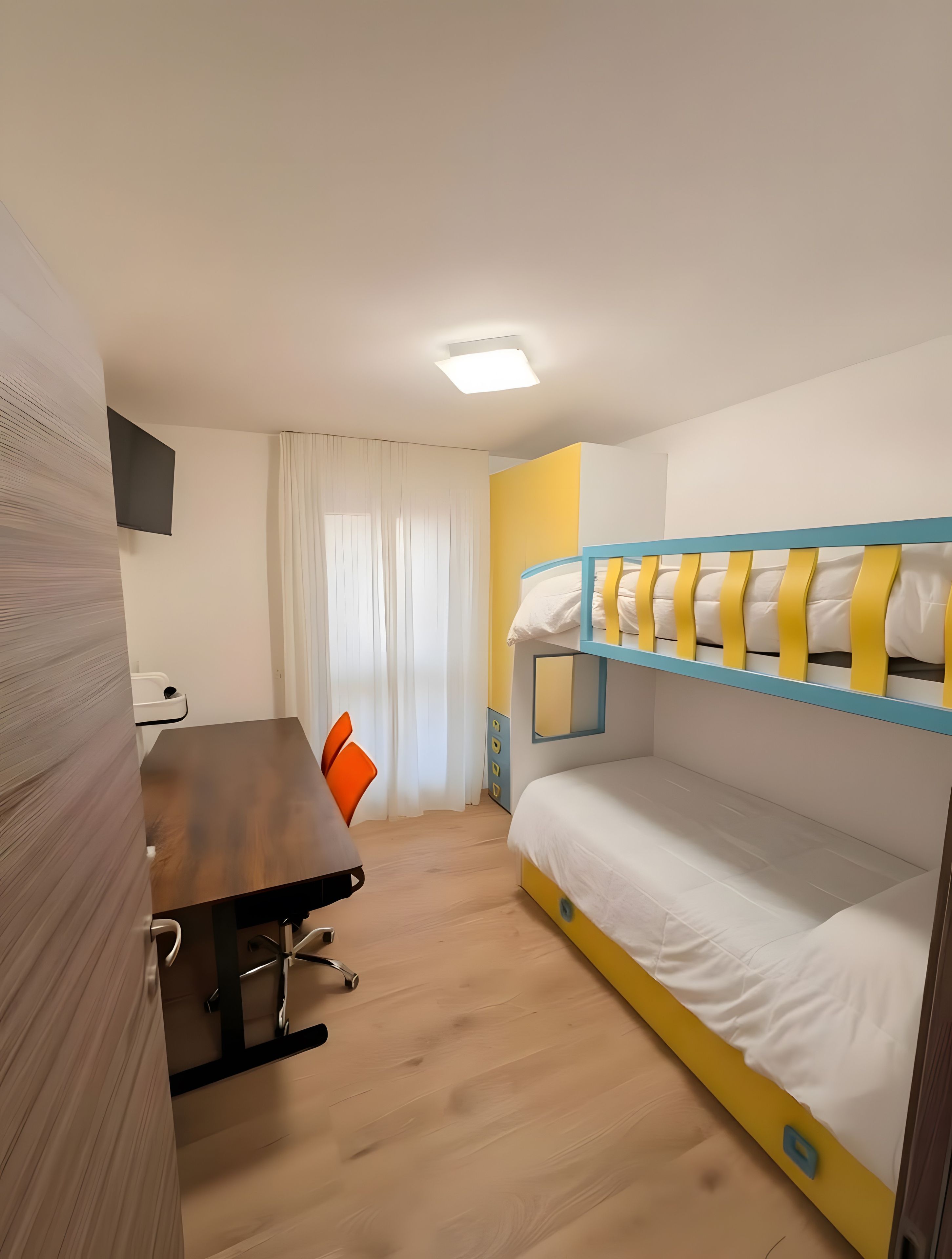 A room with a bunk bed, a desk with a chair, a window with curtains, and a flat-screen TV mounted on the wall.