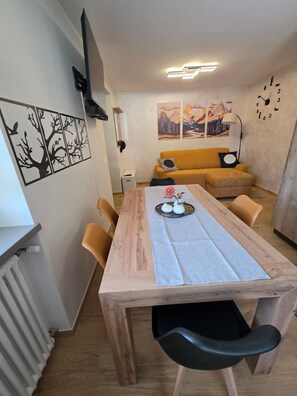 A dining area with a wooden table, chairs, and a central floral arrangement. A sofa and a lamp are visible in the background. - Alba di Poggio al Sole Apartment in the Dolomites of Val di Sole (Mezzana)