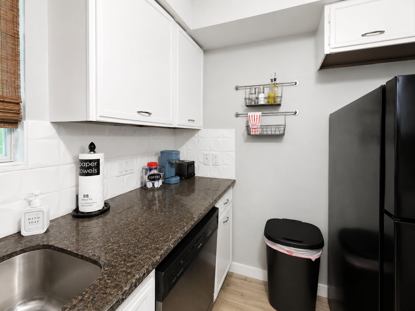 A modern kitchen with white cabinets, a black refrigerator, a coffee machine, and a trash bin.