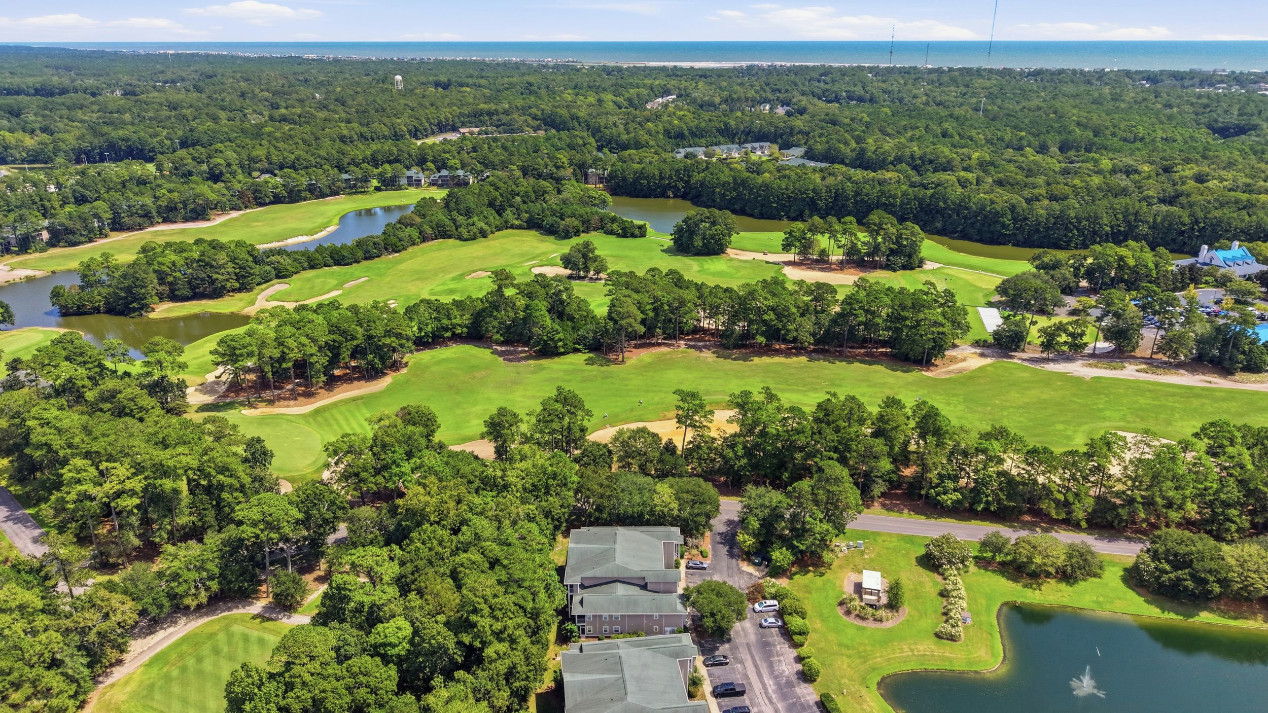 An aerial view of a golf course with multiple holes, a pond, and surrounding wooded areas.