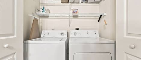 A laundry room with two front-loading washing machines and a shelf above containing various items.