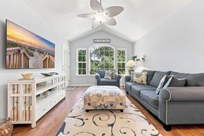 Un salon chaleureux avec une grande télévision, un canapé gris, une console en bois et une vue sur l’extérieur grâce à de grandes fenêtres.