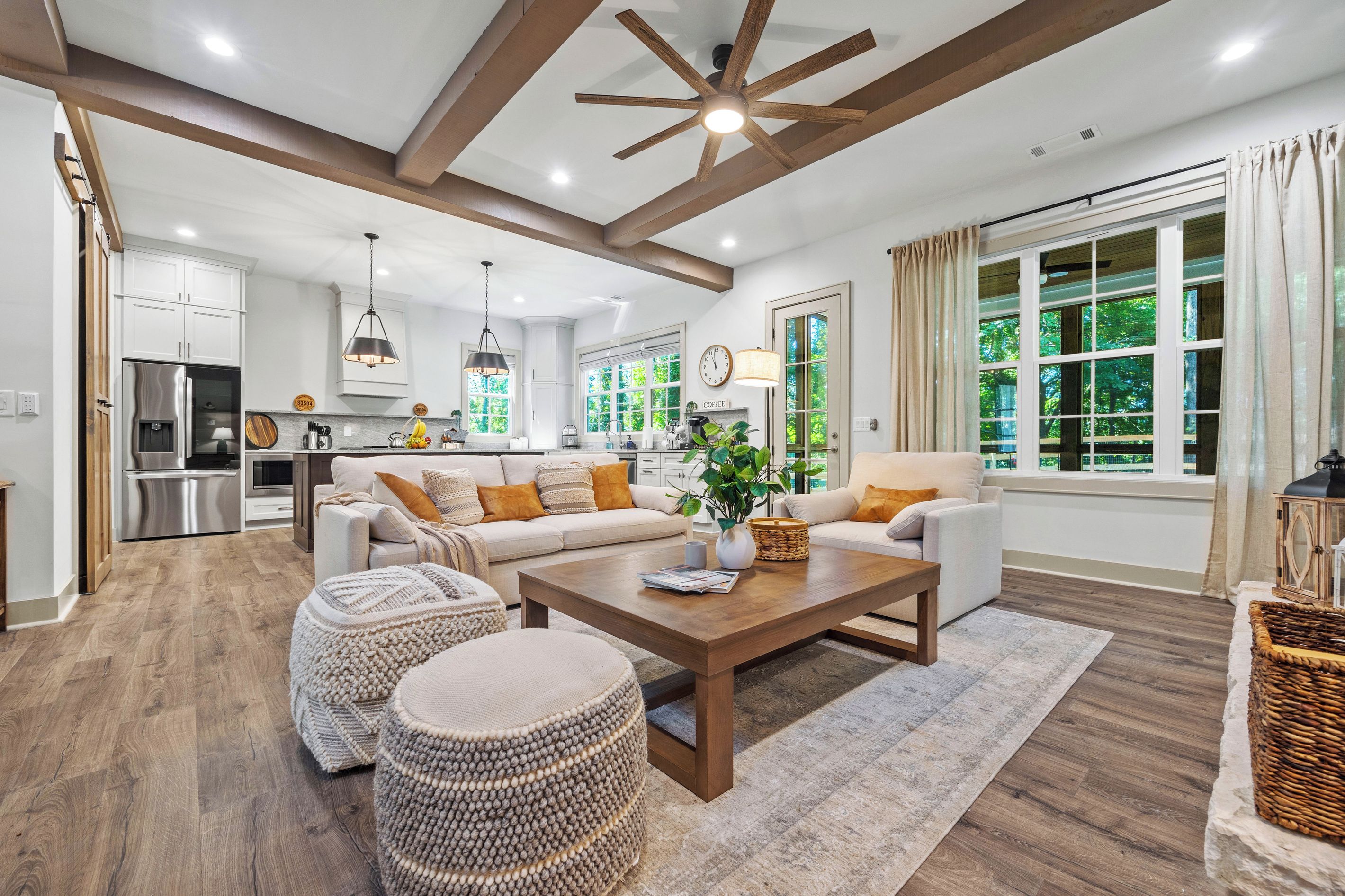 A spacious living room with a wooden ceiling, a large window with a view of greenery, and a kitchen in the background.