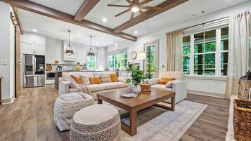 A spacious living room with a wooden ceiling, a large window with a view of greenery, and a kitchen in the background.