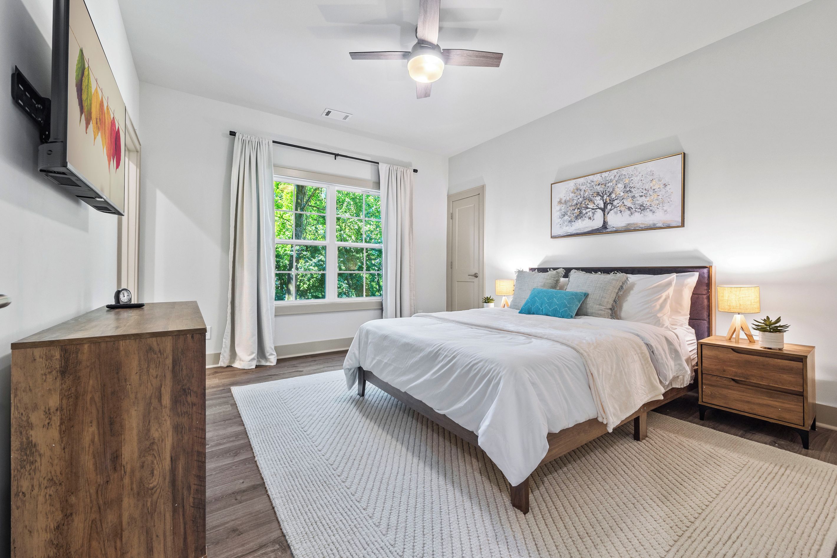 Une chambre à coucher comprenant un lit, des tables de chevet, un ventilateur de plafond, une fenêtre avec des rideaux et une œuvre d’art accrochée au mur.
