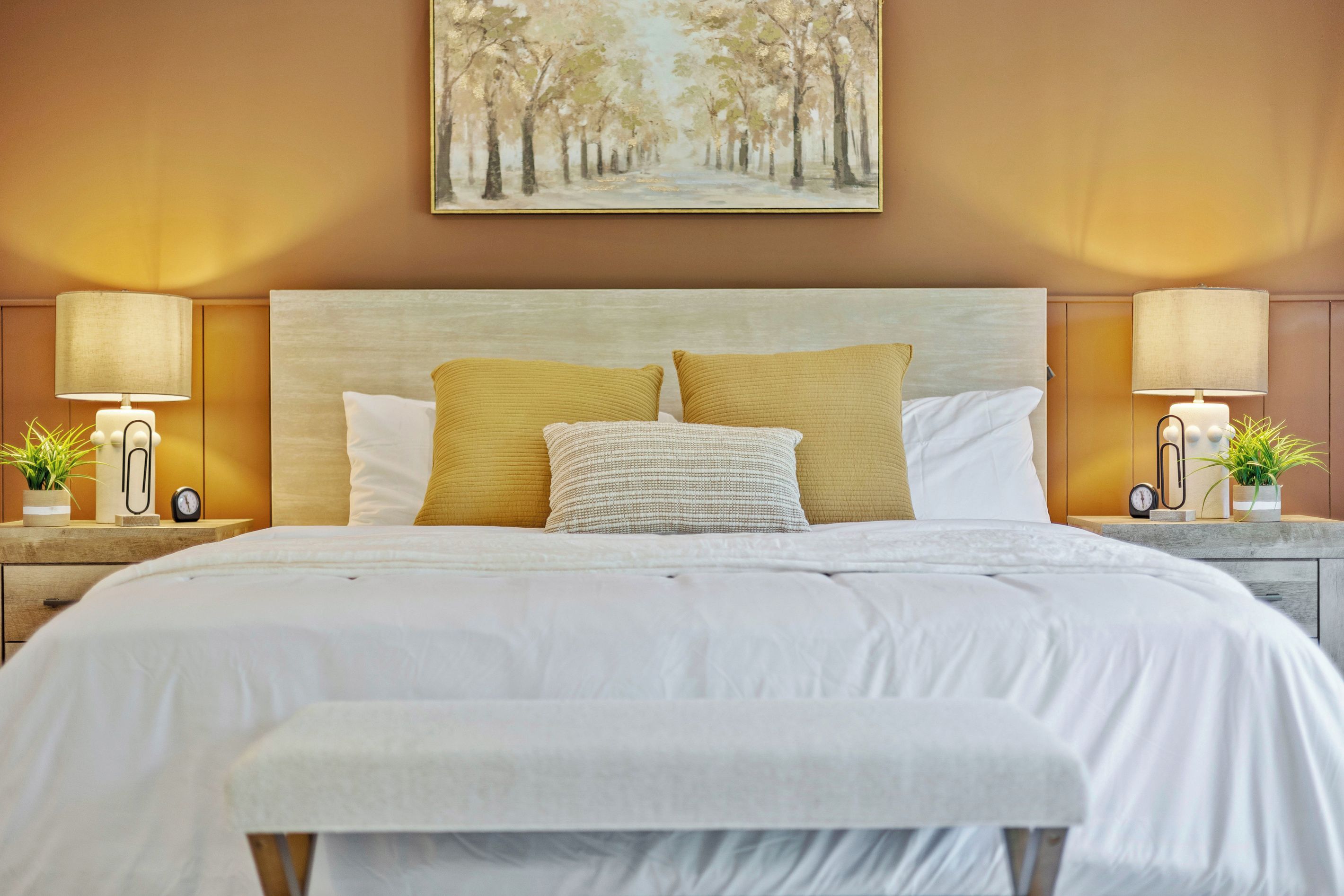Un lit bien fait, avec une literie blanche, deux oreillers jaunes et un coussin décoratif blanc. Il y a deux tables de chevet avec des lampes et des réveils, et un tableau représentant un chemin bordé d’arbres est accroché au mur au-dessus du lit.