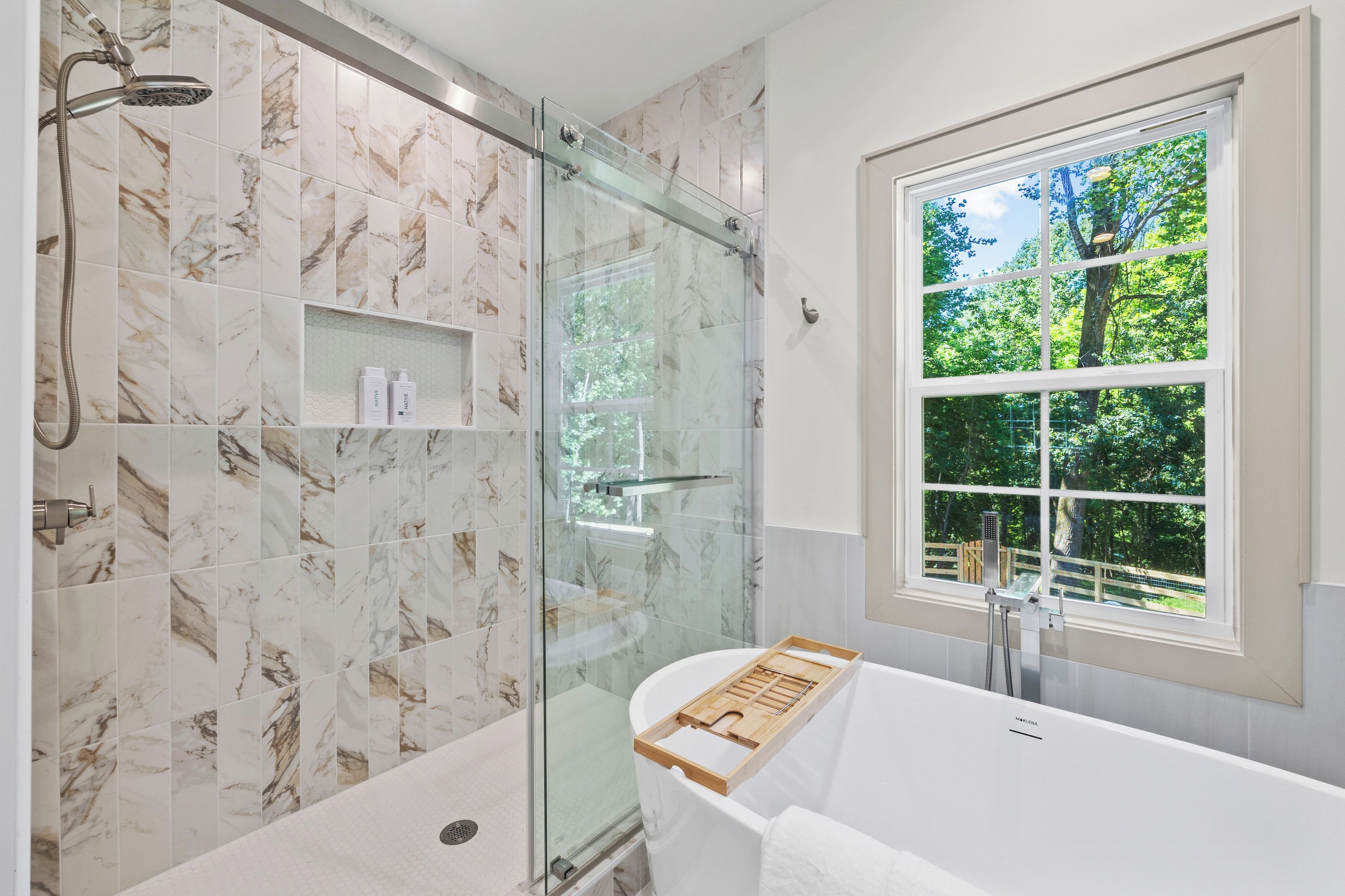 A modern bathroom with a glass-enclosed shower, a bathtub with a wooden tray, and a window with a view of trees.