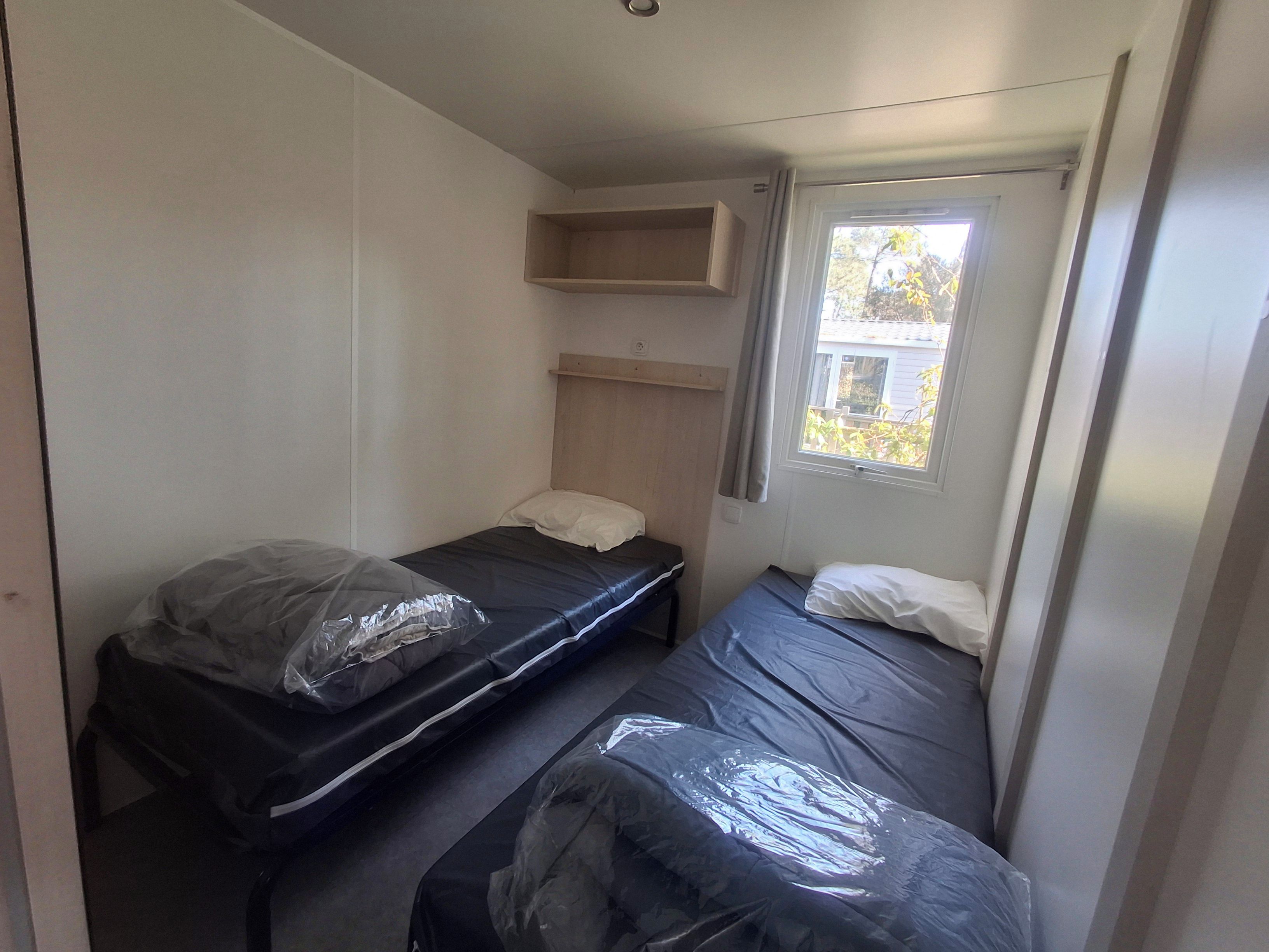 Una habitación pequeña con dos camas, un ventanal y estantería empotrada.