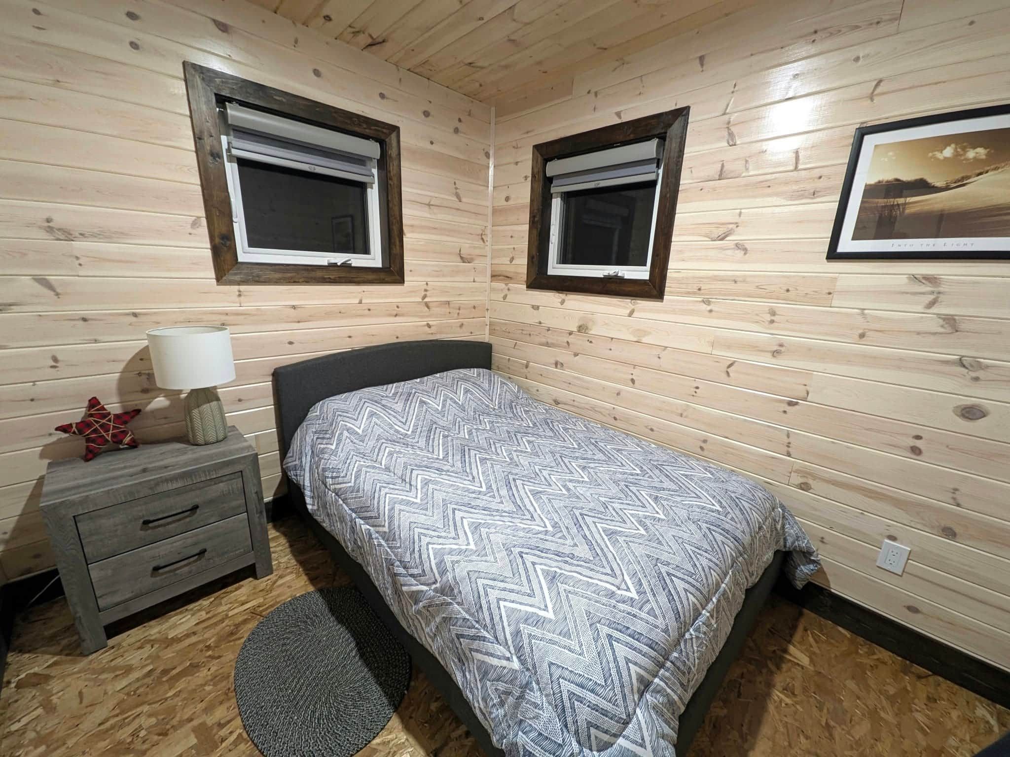 Een slaapkamer met twee ramen, een bed met beddengoed met een patroon, een nachtkastje met een lamp en een klein decoratief object.