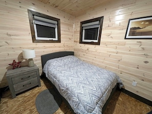 A bedroom with two windows, a bed with patterned bedding, a bedside table with a lamp, and a small decorative item.