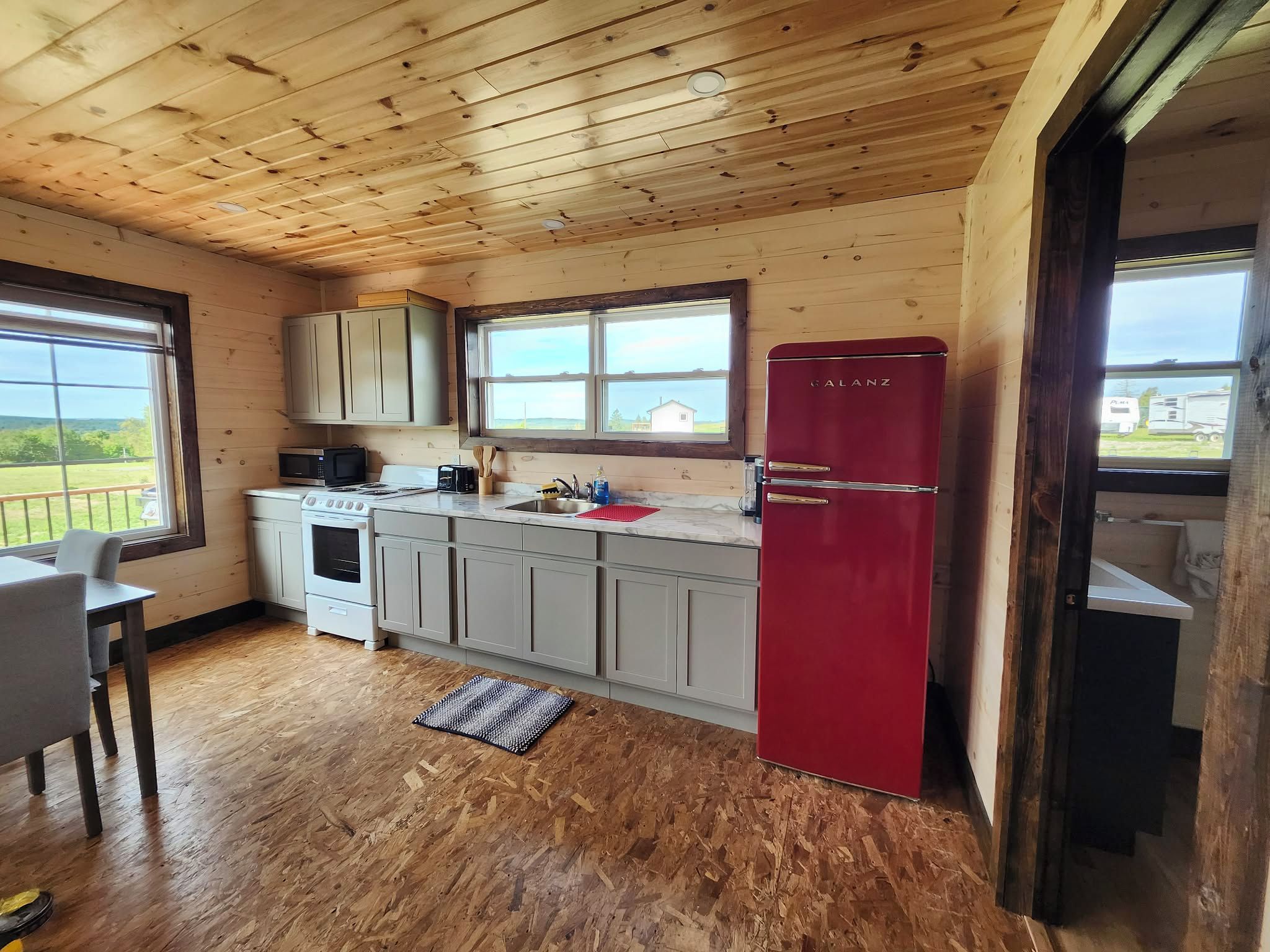 A compact kitchen with wooden ceiling, a red refrigerator, and a window overlooking a rural landscape.